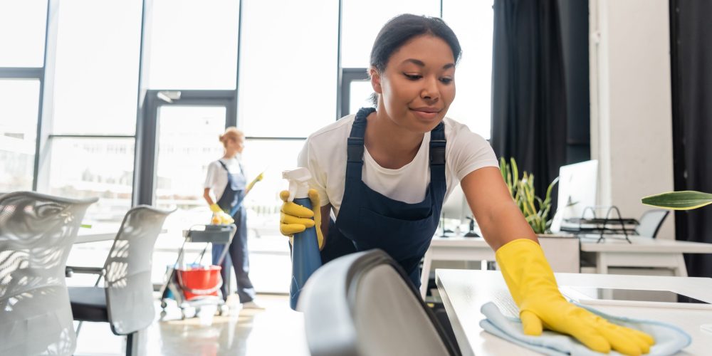 Smiling,Bi-racial,Woman,Cleaning,Office,Desk,With,Rag,Near,Colleague